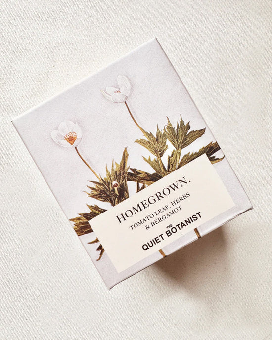 A box labeled Home Grown Candle by The Quiet Botanist, featuring tomato leaf, herbs & bergamot scents. Made with natural coconut wax and decorated with botanical illustrations on a light background.