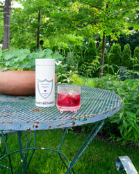 A tin of The Quiet Botanist Iced Mint Tea and a glass of its red, caffeine-free drink sit on a weathered metal table in a lush green garden.