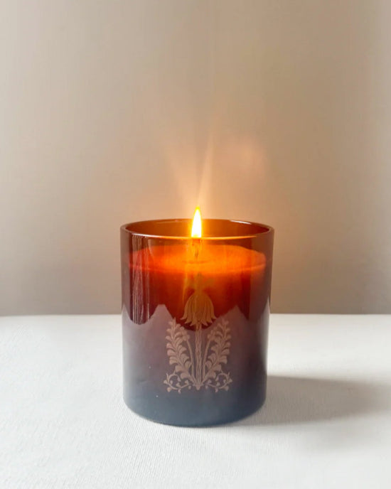 The Quiet Botanists In The Weeds candle, hand-poured into a brown glass holder with a decorative pattern, is displayed on a white surface against a plain background.