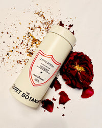 A white cylindrical container labeled Love Poem Tea by The Quiet Botanist rests on a white surface, surrounded by dried tea leaves and a dried red flower, evoking a soothing, caffeine-free turmeric infusion.