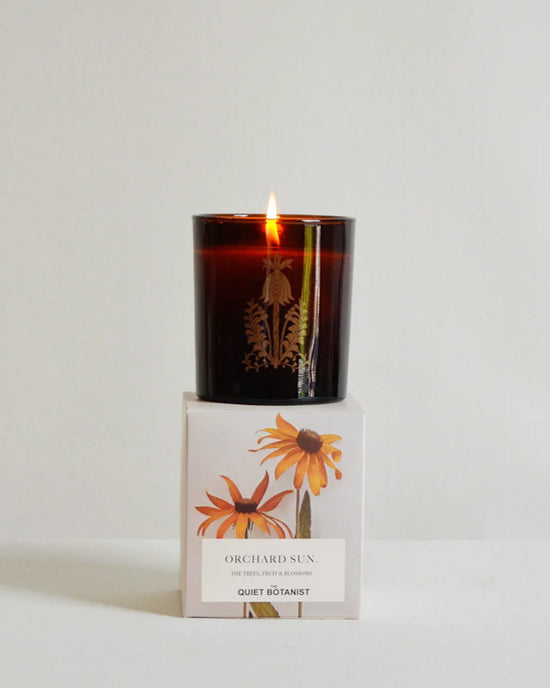 A lit brown glass Orchard Sun Candle by The Quiet Botanist rests on a white box with yellow flowers. Made from natural coconut wax and hand-poured, it adds a gentle glow to any space.