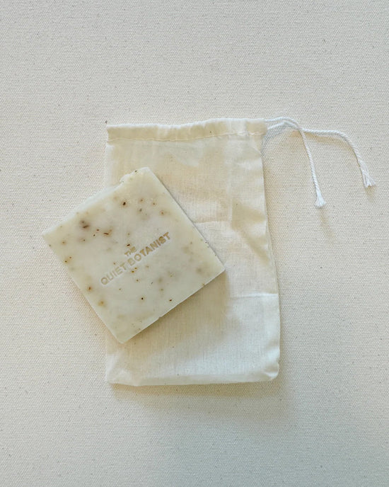 A square bar of Smell The Roses - Handmade Soap by The Quiet Botanist sits atop a white drawstring cloth bag, displayed on a light background.