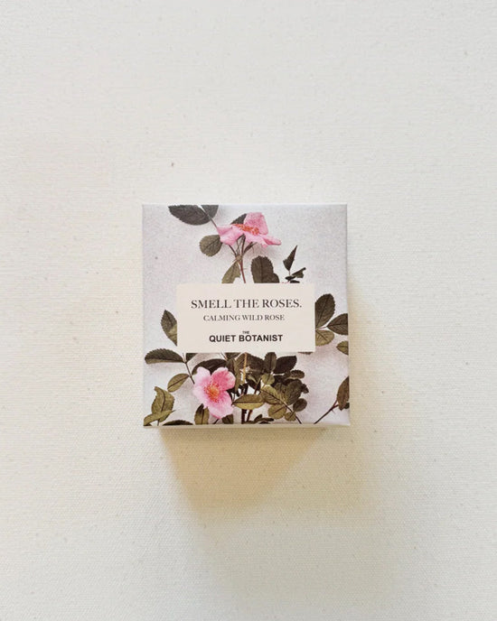 A small box labeled Smell The Roses - Handmade Soap by The Quiet Botanist, decorated with pink wild roses and green leaves on a light background, contains natural soap.