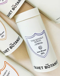 White cylindrical containers labeled THE QUIET BOTANIST feature The Kindly Moon Sleep Tea, a naturally caffeine-free blend, arranged on a light surface with shield designs and botanical branding.