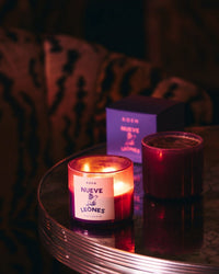 A lit Nueve Leones Candle by ROEN, made from a coconut soy wax blend and premium fragrance oils, sits on a round reflective table beside an unlit candle and purple box, with a patterned chair in the dim background.