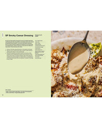 A spoon drizzles creamy Caesar dressing over salad with cheese, chicken, and greens. Find The Salad Project by Random House on the left—your go-to for salad inspiration and the perfect addition to your next cookbook creation.