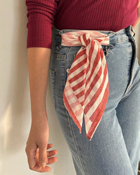 Someone in blue jeans and a red long-sleeve top wears a Casa Amarosa Stripe Hand Block Printed Scarf in Red & Pink, tied in a bow at their waistband, while their left arm hangs relaxed at their side.
