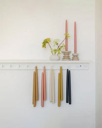 A white wall shelf displays two pink hand-dipped candles in holders and a small vase with green and brown leaves. Below, The Floral Society 12in Beeswax Taper Candles hang from pegs in tan, pink, gold, and black.