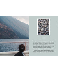 A person with dark hair gazes from a boat at a rocky mountain by calm water—a scene from Travel Guide: Somewhere in Italy by Cozy Publishing. On the right, round gray stones are shown on a pale blue page with text below.