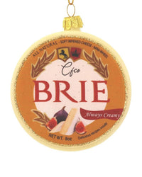 The Cody Foster Brie Ornament is a hand-crafted round glass decoration resembling a wheel of brie, featuring a BRIE label, cheese and fig illustrations, gold accents, and hangs by a shimmering gold string.