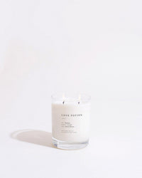 The Brooklyn Candle Studio Love Potion Classic 2-Wick Candle, made from soy wax, burns in a clear glass jar labeled LOVE POTION on a white background, with the jar casting a faint shadow.