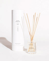 A glass Brooklyn Candle Studio Santa Fe Reed Diffuser with wooden sticks sits next to a tall white Room Perfume box, both on a plain light background.