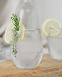 A Pebbled Spirits Glass by The Collective, filled with ice water and garnished with lemon and rosemary, sits on wood. Its recycled glass subtly pebbled; a matching glass from the set of 4 is visible in the background.