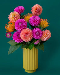 A Monroe Vase in Palm by Jungle Club holds pink, orange, and yellow dahlias with green leaves, set against a dark green background.