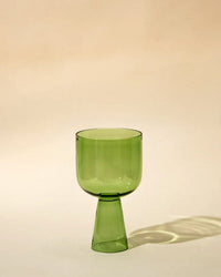The Casa Amarosa Aria Glass in Green features a modern geometric design with a wide, tapered base. This pastel goblet sits on a neutral beige background, casting a subtle shadow.