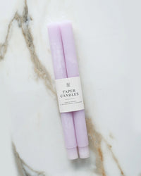 Two hand-dyed Lavender Taper Candles by BOTANICA, wrapped in a white label reading "Taper Candles," are displayed on a white marble surface with brown and gray veining.