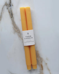 Two BOTANICA Goldenrod Taper Candles | Fluted are hand-dyed in yellow and wrapped in a white label reading "Taper Candles," displayed on white marble with gray and brown veins.