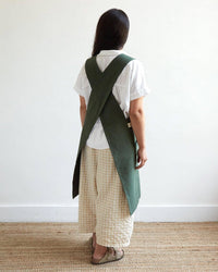 A person stands facing away, dressed in a white shirt, beige checkered pants, sandals, and The Floral Society's Canvas Workshop Apron in Fern. The simple room features a woven rug and a white wall.