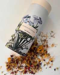 A cylindrical Edible Flower Blend container by The Quiet Botanist, adorned with botanical illustrations, lies on its side as vibrant dried flowers spill out—ideal for cake decorating—across a light surface.