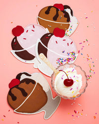 Four vibrant Ice Cream Sundae Napkins by Piecework Puzzles are arranged on a pink background with sprinkles. A real Neapolitan sundae topped with whipped cream, cherry, and more sprinkles adds a playful touch in the corner.