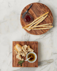 The etúHOME New Classic Round Trivet Riser, Medium displays breadsticks, a jam jar, and a knife above a square acacia wood trivet with torn bread, oil bowl, spoon, and rosemary on marble.