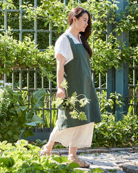 A woman in The Floral Society’s Canvas Workshop Apron in Fern walks barefoot on a garden stone path, holding greenery. She wears a light dress; lush plants and a metal fence are visible in the background.