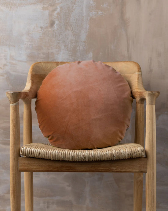 A wooden chair with a woven seat displays the Saarde Linen/Velvet Round Pillow with Feather Insert in Terracotta, set against a textured beige wall.