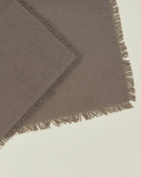 Two overlapping dark grey Essential Cocktail Napkins from Hawkins New York, made of 100% cotton with frayed edges, are arranged diagonally on a cream surface. Set includes 4 napkins.