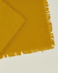 A set of four Essential Cocktail Napkins in Mustard by Hawkins New York, made from 100% cotton with frayed edges, is displayed on a light surface with one napkin partially overlapping another at an angle.