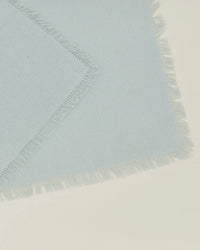 A close-up of two overlapping Hawkins New York Essential Cocktail Napkins in Sky—100% cotton with fringed edges—resting on a cream-colored surface.