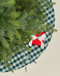 A top-down view of a Christmas tree with green branches stands on the Hawkins New York Essential Gingham Tree Skirt in Sky/Peacock. A red gift box with a white ribbon lies near the edge of the tree skirt.