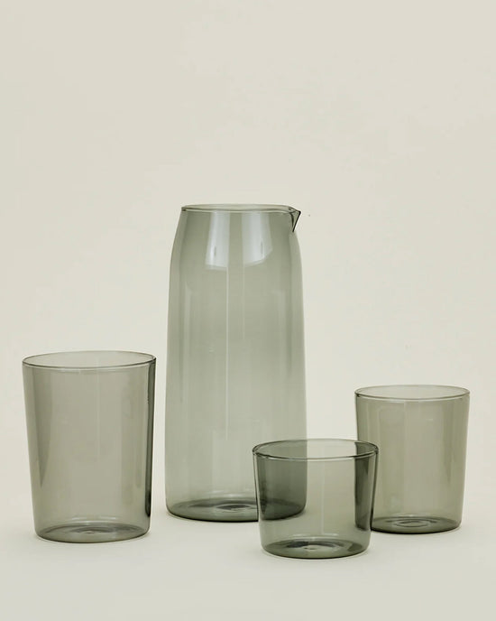 A set of Hawkins New York Essential Glassware—Pitcher in Smoke with a spout and three matching smoky gray tumblers of different heights, showcased on a light background.