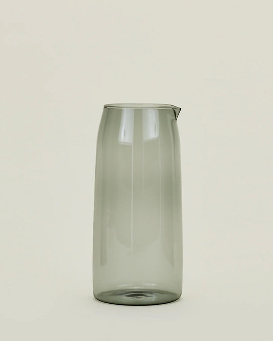 A tall, transparent 52 oz borosilicate glass pitcher in Smoke by Hawkins New York stands upright against a light beige background, featuring a small spout and exuding simplicity and modern elegance.