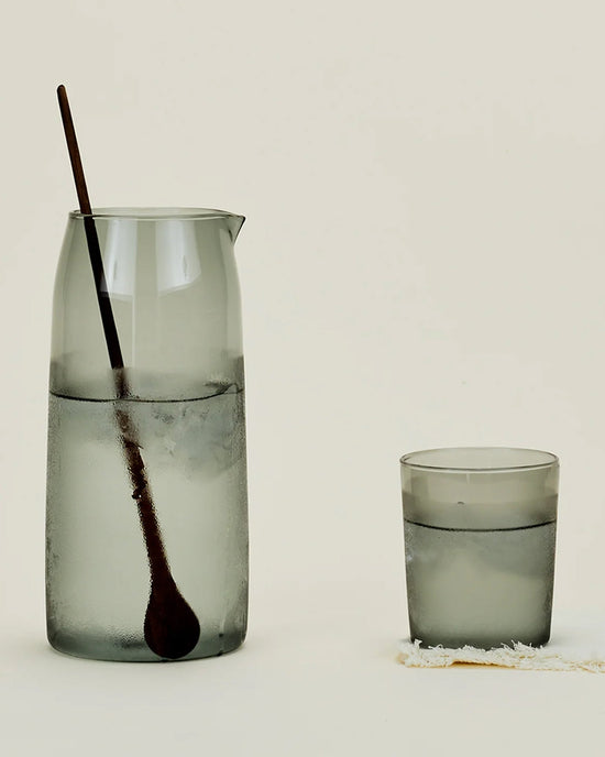The Essential Glassware - Pitcher in Smoke from Hawkins New York, a frosted 52 oz borosilicate glass pitcher with wooden spoon, stands next to a matching water glass on a light surface with visible condensation.