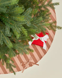 A top-down view of a Christmas tree with green branches, a red gift with a white ribbon, and the Hawkins New York Essential Striped Tree Skirt in Blush/Terracotta tied with a bow.