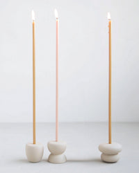 Three tall, thin candles—two beige and one light pink—are lit and standing upright in The Floral Society's Bulb Petite Long Stem Candleholders in Sand, set against a plain white background.