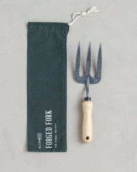 The Floral Society’s Forged Fork, crafted from durable boron steel with a light wooden handle, rests beside its dark green, white-drawstring pouch labeled "Forged Fork" on a light gray surface.