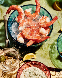 A blue bowl of ice and large shrimp, inspired by Cook This Book from Random House, sits on a green marbled surface with a fork, iced drink, lemon wedges, and a red bowl of creamy dipping sauce.