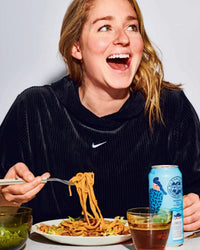 A smiling woman in a dark hoodie eats noodles with chopsticks at a table, using easy recipes from Cook This Book by Random House, with a can of sparkling water and a glass of tea beside her.