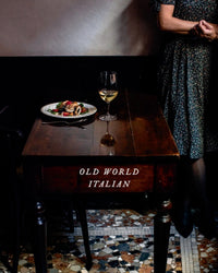 A wooden table set with Italian food and white wine. The table bears the words "Old World Italian" by Random House. A woman in a floral dress stands partially visible, capturing the warmth of authentic Italian recipes.