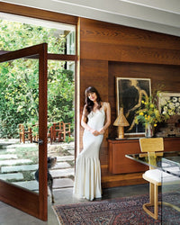 A woman in a long white dress stands with her dog in a chic, wood-paneled room from Ad At Home: Architectural Digest by Random House—modern art and flowers indoors, with a lush garden and wooden dining set visible outside.
