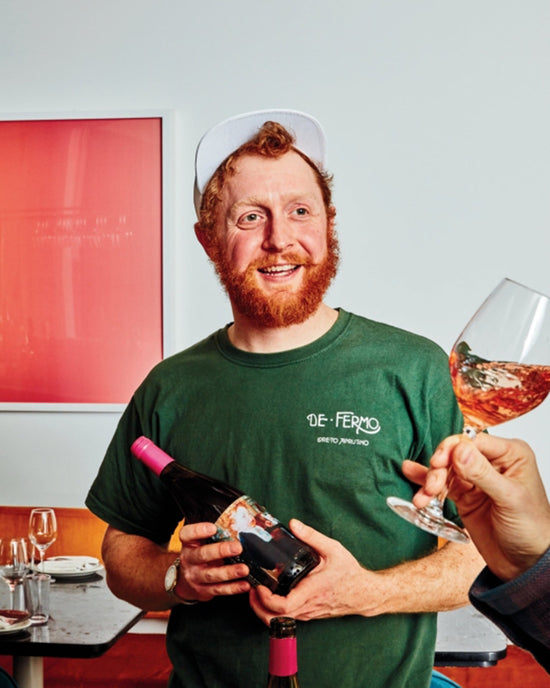 A smiling man with a red beard, wearing a green shirt and white cap, holds a bottle of Salad Pizza Wine by Random House. Another hand in the foreground raises a glass of rosé toward him, with red art hanging on the wall behind them.