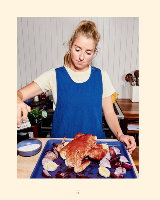 Inspired by easy recipes from Cook This Book by Random House, a woman in a blue apron pours liquid from a bottle over whole raw chicken and sliced onions on a baking tray in her kitchen.