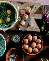 A rustic kitchen scene inspired by Old World Italian from Random House features eggs, a knife on a wooden board, spinach quiche with raw eggs, eggplants, coarse salt, and white wine on a wooden table.
