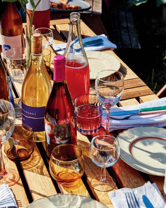 A wooden outdoor table set with plates, natural wines, rosé-filled glasses, and napkins evokes a relaxed meal inspired by Salad Pizza Wine from Random House.