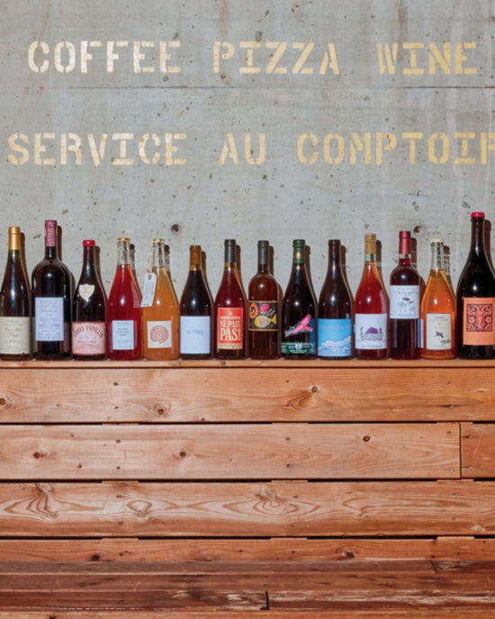 A row of colorful Salad Pizza Wine bottles by Random House stands on a wooden counter, set against a concrete wall with faded COFFEE PIZZA WINE SERVICE AU COMPTOIR text.