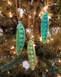 Three Garden Pea - Teal Green Ornaments by Cody Foster, hand-crafted glass pieces shaped like pea pods, hang on a Christmas tree with green needles and glowing white string lights in the background.