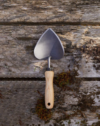 The Floral Society Potting Trowel, featuring a smooth wooden handle and boron steel blade, rests on a weathered wood surface with moss—an essential addition to your favorite garden tools.