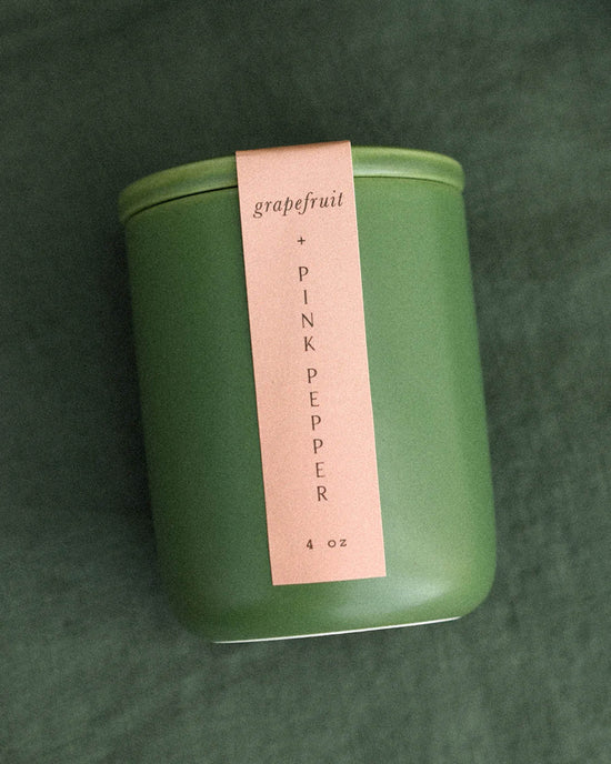 The LEIF Grapefruit & Pink Pepper Candle, a 4 oz green jar with a pink label, features a soy wax blend and wooden wick, and is shown resting on green fabric.