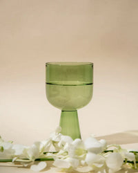 The Aria Glass in Green by Casa Amarosa stands on a light surface, filled with water and surrounded by white flower petals against a beige background—showcasing pastel glassware elegance.